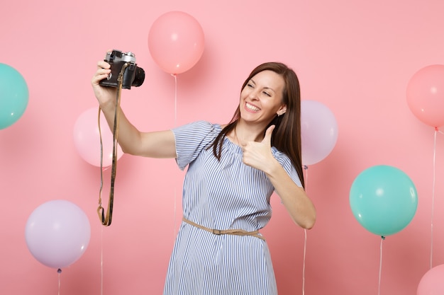 Portrait of joyful young woman in blue dress doing selfie on retro vintage photo camera showing thumb up on pink background with colorful air balloons. Birthday holiday party people sincere emotions.