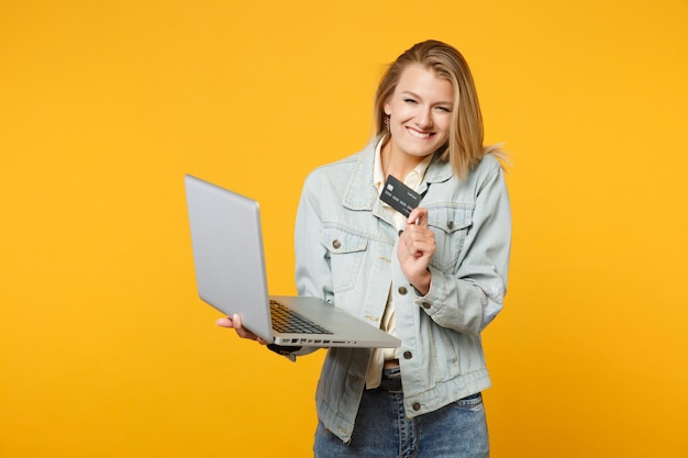 Portrait of joyful young woman in denim casual clothes holding laptop pc computer, credit bank card isolated on yellow orange wall background in studio. People lifestyle concept. Mock up copy space.