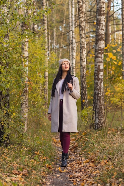 Portrait of a joyful young woman enjoying in the autumn park. Beautiful brunette girl in autumn grey coat and knitted hat. Relax in nature.