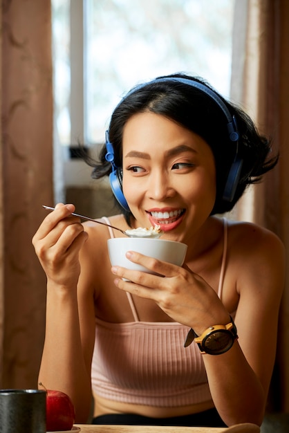 Ritratto di una giovane donna gioiosa che ascolta musica in cuffia e mangia un delizioso muesli con yogurt