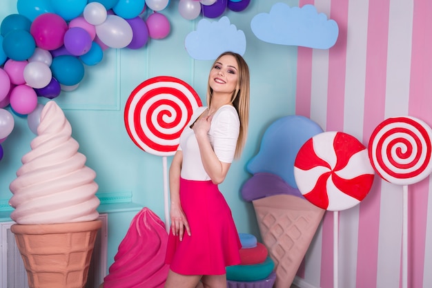 Portrait of joyful young woman  in pink dress on background decorated with huge candies and ice cream.