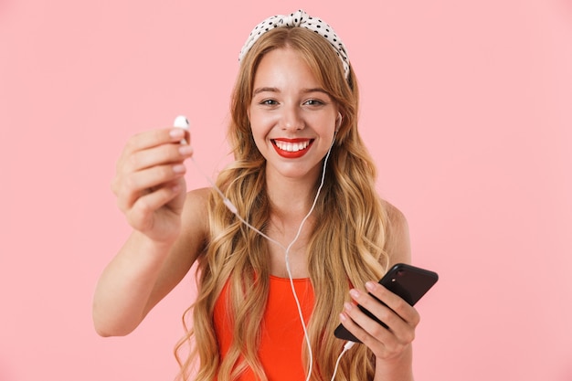 Portrait of joyful young woman with long curly hair smiling and holding smartphone with earphones isolated over pink wall