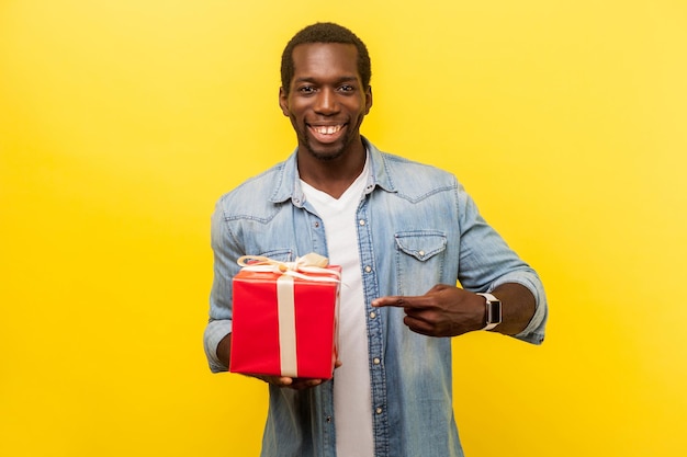 Portrait of joyous handsome man in denim casual shirt smiling at camera and pointing at red gift box satisfied with present enjoying holidays indoor studio shot isolated on yellow background