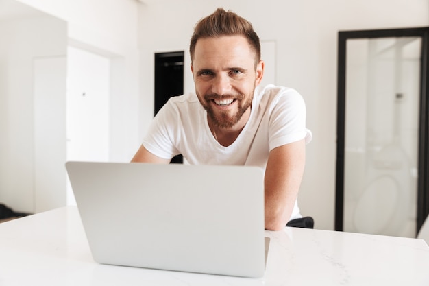 Portrait of joyous satisfied man smiling, while sitting at table and using silver laptop in white bright room