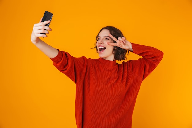 Портрет радостной женщины 20-х годов в свитере, держащей и делающей селфи-портрет на мобильном телефоне, стоя изолированной над желтым