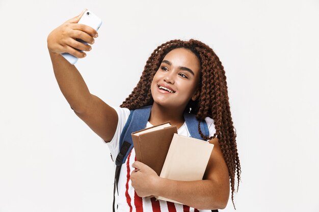 Ritratto di una donna gioiosa che indossa uno zaino che scatta una foto selfie sul cellulare mentre tiene in mano libri che studiano isolati contro un muro bianco