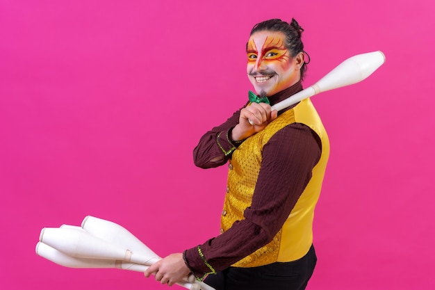 Photo portrait of a juggler in a waistcoat and with painted face juggling maces on a pink background