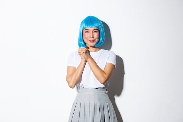 Portrait of kawaii asian girl looking grateful and amused, clasp hands together and smiling pleased at camera, standing in blue wig and schoolgirl costume for halloween party.