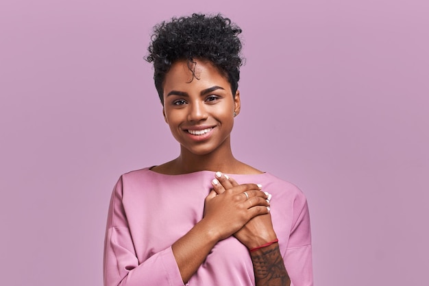 Photo portrait of kind hearted african american female in fashionable overalls, keeps hands on chest, shows her kindness and sympathy, has pleased cheerful expression, isolated over lavender background