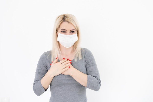 Portrait of kind hearted female wearing a respiratory mask from coronavirus disease, keeps hands on chest, shows her kindness and sympathy, on beige wall. COVID-19 epidemic.