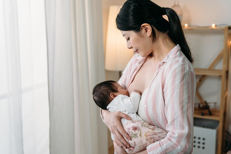 Photo 肖像画 窓の近くに立っている韓国の初めてのお母さんは、明るい家のインテリアで彼女に母乳を与えながら、愛情を込めて揺りかごにいる赤ん坊の娘を見つめています。