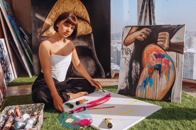 Portrait of a lady artist sitting on a carpet in an art studio with a view of the city among portraits and painting equipment