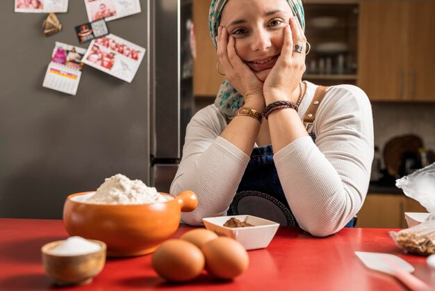 Портрет латинской взрослой женщины-повара, сидящей на кухне с руками на щеках, улыбающейся и смотрящей в камеру