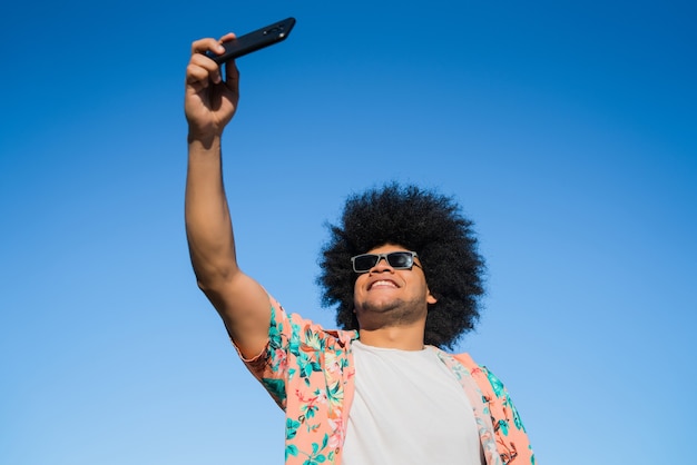 Ritratto di uomo latino prendendo un selfie con il suo telefono cellulare stando in piedi all'aperto sulla strada