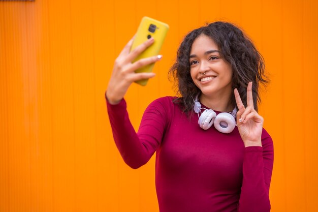 Portrait of latin woman with headphones smiling technology concept on an orange background taking a selfie