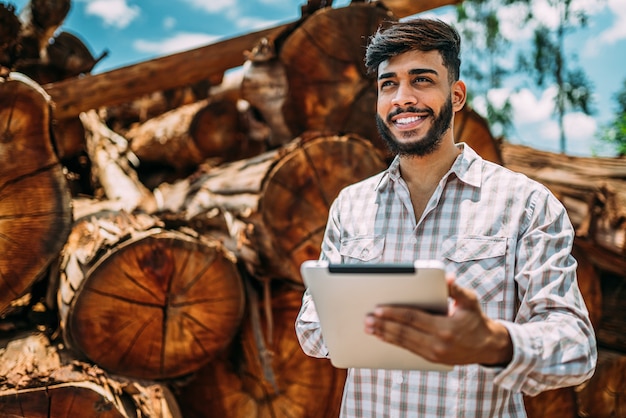 Portrait of Latin young man working on tablet beside tree trunks.