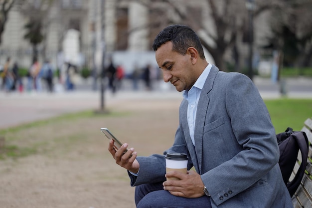 Portrait of latino man in jacket looking a mobile phone sitting on a bench in a public park