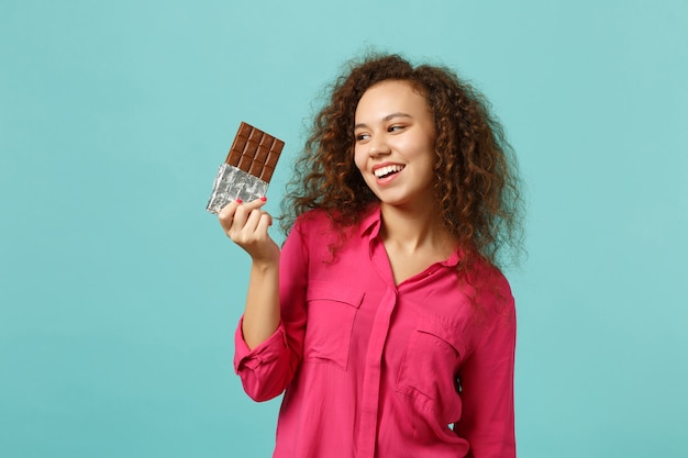 Portrait of laughing african girl in casual clothes holding in hand chocolate bar isolated on blue turquoise wall background in studio. People sincere emotions, lifestyle concept. Mock up copy space.