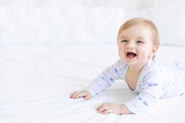 Photo portrait of a laughing baby boy blonde lying in a crib on his stomach in a room at home with blue and white cotton bedding and smiling in the morning the concept of children's goods and accessories