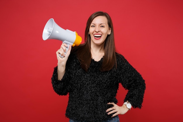 Portrait of laughing cheerful cute young woman in black fur sweater holding megaphone isolated on bright red wall background in studio. People sincere emotions, lifestyle concept. Mock up copy space.