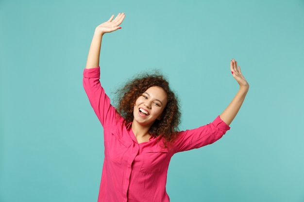 Portrait of laughing cheerful pretty african girl in casual clothes rising hands isolated on blue turquoise wall background in studio. People sincere emotions, lifestyle concept. Mock up copy space.