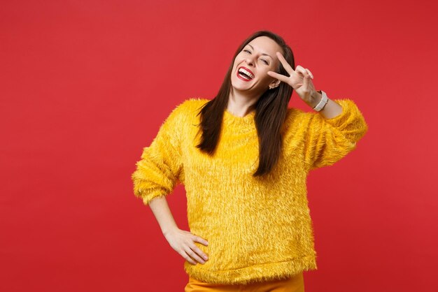 Portrait of laughing funny young woman in yellow fur sweater showing victory sign isolated on bright red wall background in studio. People sincere emotions, lifestyle concept. Mock up copy space.