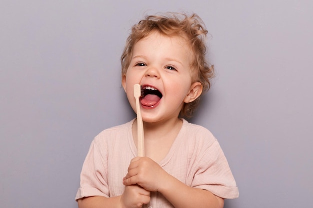 Portrait of laughing happy cheerful infant girl kid with curly blonde hair brushing her teeth taking care of dental hygiene standing isolated over gray background