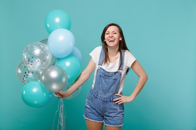 Portrait of laughing happy cute young woman in denim clothes celebrating and holding colorful air balloons isolated on blue turquoise wall background. Birthday holiday party, people emotions concept.