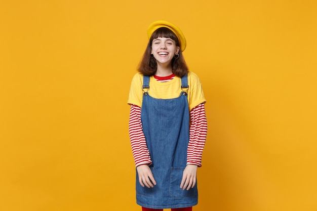 Portrait of laughing joyful bright girl teenager in french beret, denim sundress standing isolated on yellow wall background in studio. people sincere emotions, lifestyle concept. mock up copy space.