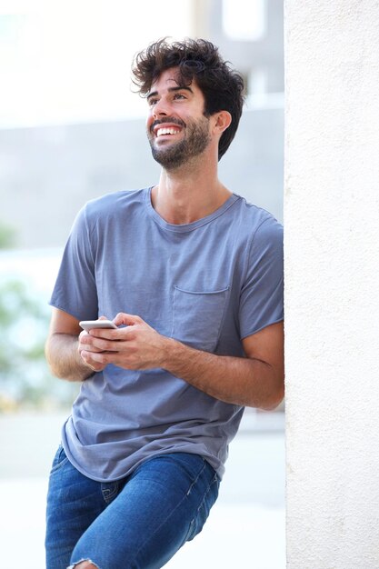 Portrait of laughing man holding phone leaning on wall outside