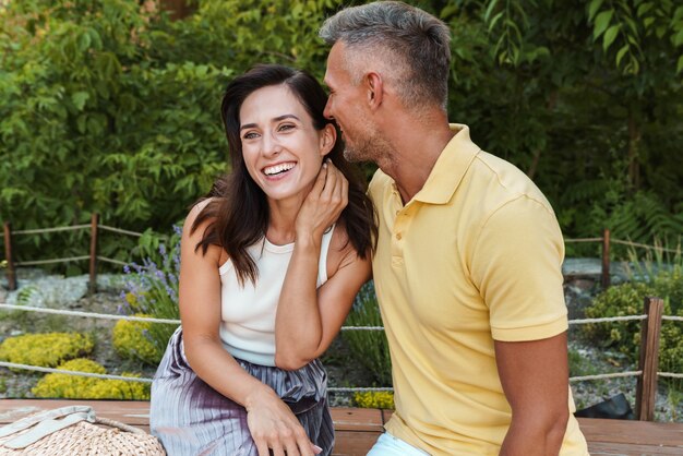 Portrait of laughing middle-aged couple man and woman hugging while sitting on bench in summer park