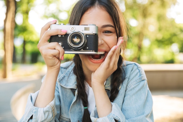 Ritratto di una giovane studentessa carina felice ottimista ridente che indossa occhiali seduto su una panchina all'aperto nel parco naturale che tiene la fotocamera