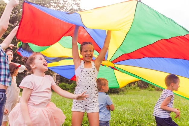 Portrait of laughing preteen girl supporting large colorful canopy holding her hands up while group of happy children running under it Support and care of younger ones concept