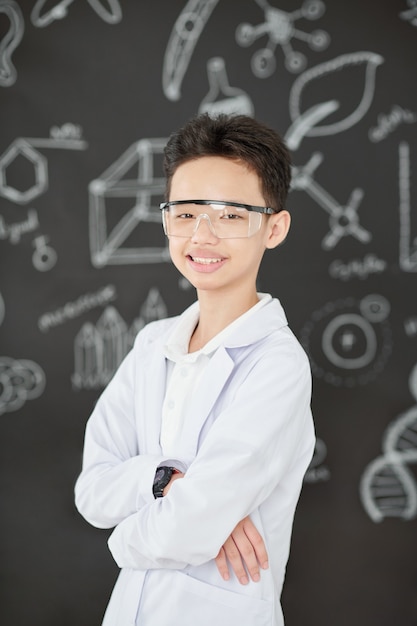 Portrait of laughing schoolboy in labcoat standing against balckboard with scientific drawings