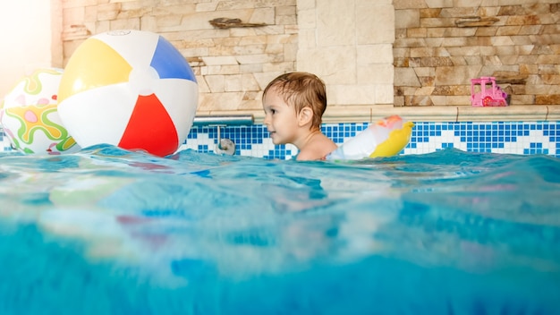 Portrait of laughing and smiling 3 years old little boy swimming with inflatable colorful ring and playing with beach ball in swimming pool