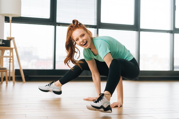 Portrait of laughing sporty young woman in sportwear doing advanced yoga pose at home on background of window looking at camera