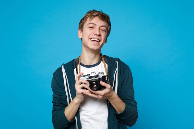 Portrait of laughing young man in casual clothes holding in hands retro vintage photo camera isolated on blue wall . people sincere emotions, lifestyle concept