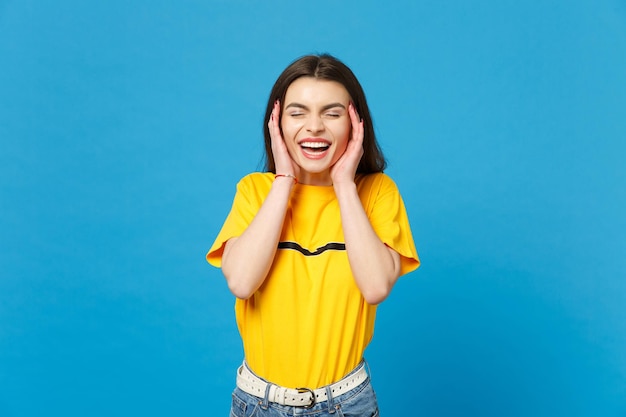 Portrait of laughing young woman in vivid casual clothes keeping eyes closed, putting hands nears face isolated on bright blue wall background in studio. People lifestyle concept. Mock up copy space.