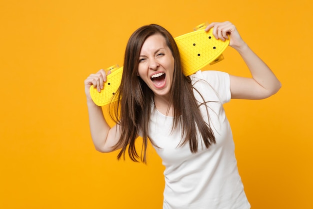 Portrait of laughing young woman in white casual clothes looking camera, holding yellow skateboard isolated on bright yellow orange background in studio. People lifestyle concept. Mock up copy space.