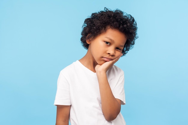 Portrait of lazy bored preschool boy with curly hair leaning on hand and looking depressed, child with indifferent gloomy expression has no energy. indoor studio shot isolated on blue background