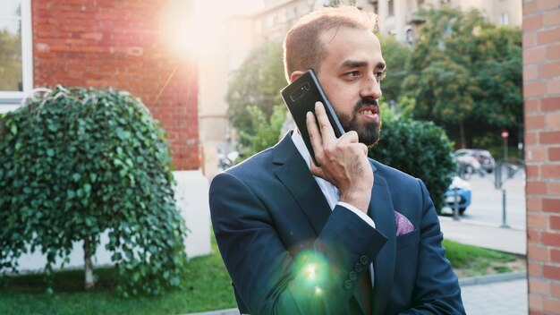 Portrait of leader man in diplomatic suit talking at phone discussing financial strategy with remote collegue. Executive manager standing outside in front of startup building office