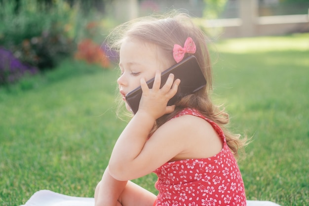 Portrait of little 3-4 girl in red clothes sitting on blanket on green grass and speaking on cell phone. Children using gadgets