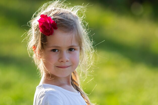 Foto ritratto di piccola ragazza bionda adorabile con gli occhi grigi e la rosa rossa in bei capelli lunghi che sorridono a porte chiuse su verde vago luminoso all'aperto