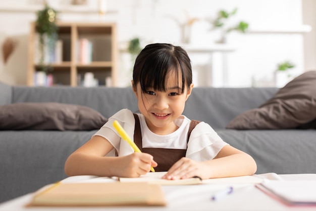 어린 아시안 소녀의 초상화: 아이가 집에서 공부하고, 미소 짓고, 행복합니다. 예쁜 소녀는 노트북에서 그림을 그리고 손으로 글을 그리며 자신의 기술과 감정적 인 이언트를 발전시니다.
