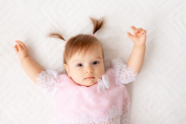 Портрет маленькой девочки шести месяцев на белой кровати в розовой одежде с поднятыми руками
