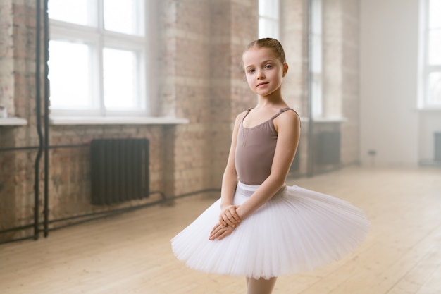 Portrait of little ballet dancer in tutu dress looking at camera standing in dance school