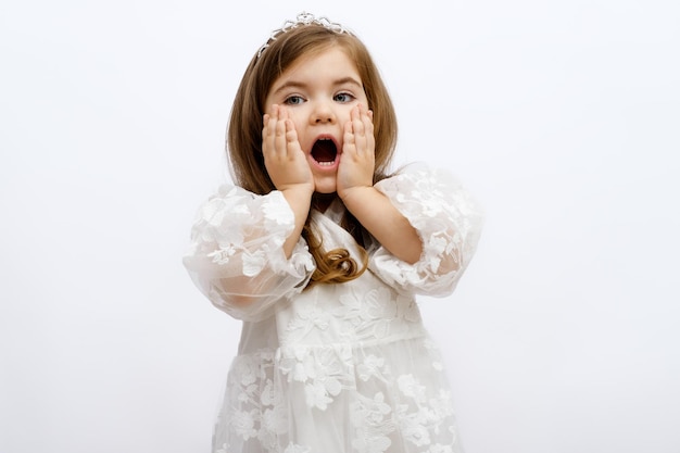 Portrait little beautiful blonde girl princess in white dress on white background with mouth open in surprise for children's advertising