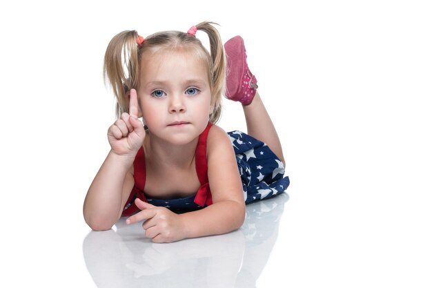 Portrait of a little beautiful girl in a dress lying on the floor with a raised index finger isolated on a white background