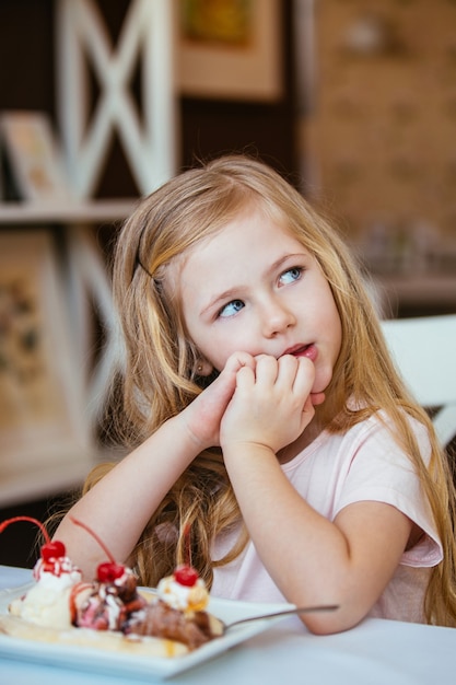 Портрет маленькой красивой девушки, сидящей в кафе за столом, мечтает с шариком мороженого с фруктами.
