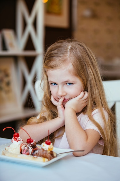 Portrait of a little beautiful girl sitting in a cafe at a table with a dish of ice cream with fruit.